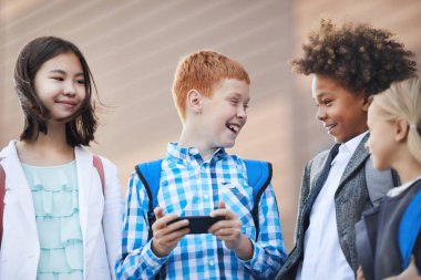 Red haired boy standing with mobile phone and laughing while talking to classmates outdoors