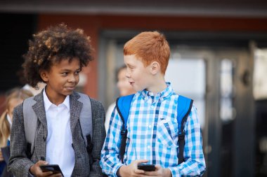 Two schoolboys with mobile phones talking to each other while walking outdoors after school lessons