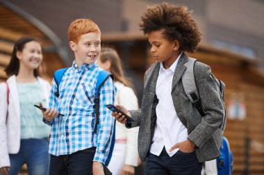 Two friends using smartphones while walking along the street after school with classmates in the background