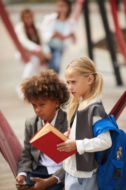 Blond schoolgirl with backpack behind back holding book standing on the playground with African boy using mobile phone