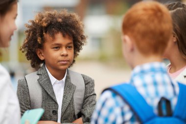 African schoolboy with curly hair standing and talking to friends outdoors after school lessons