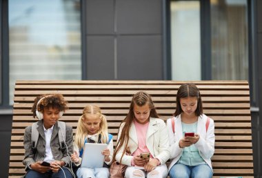 Group of school children sitting on the bench and using different gadgets studying online