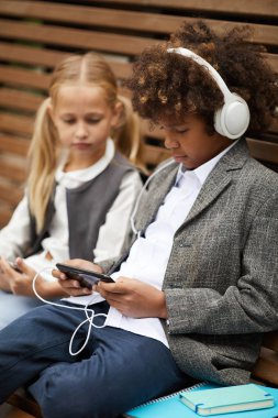 African schoolboy in headphones using mobile phone and listening to music while sitting on the bench together with girl outdoors