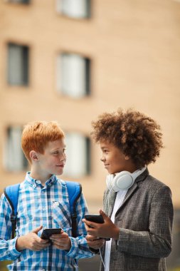 Two smiling boys standing and talking to each other and using smartphones outdoors