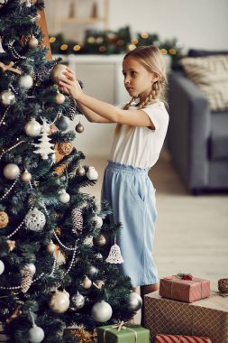 Sarı saçlı küçük kız ayakta duruyor ve Noel ağacını oyuncaklarla süslüyor.