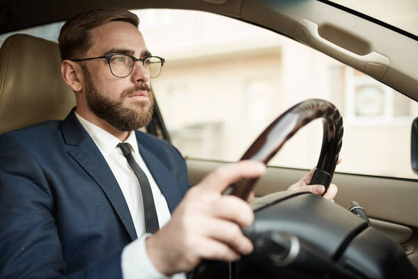 Молодой успешный бизнесмен в очках и костюме, сидящий в роскошной машине и за рулем 