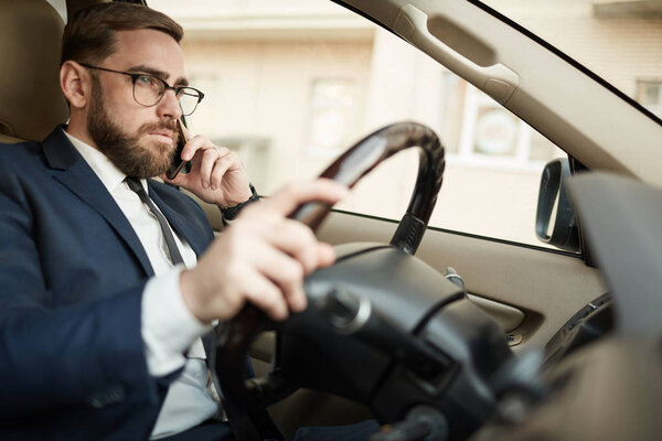 Молодой бизнесмен в очках разговаривает по мобильному телефону и водит машину 