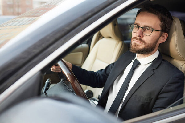 Молодой босоногий бизнесмен в очках сидит за рулем и управляет машиной, сосредоточенной на дороге