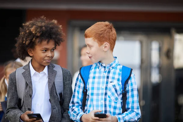 Two schoolboys with mobile phones talking to each other while walking outdoors after school lessons