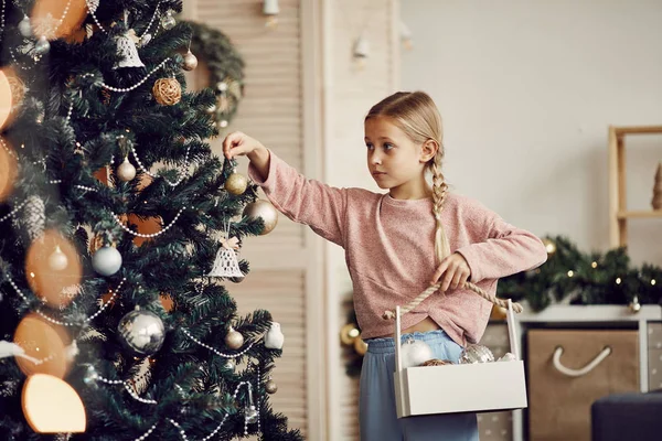 小さな女の子とともにブロンド髪保持ボックスとともにおもちゃで手とハングオンクリスマスツリー前に休日自宅 — ストック写真