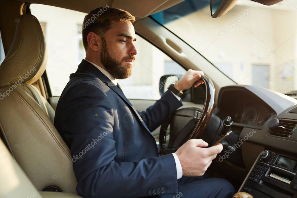 Young businessman sitting behind the steering wheel and driving while reading message on mobile phone