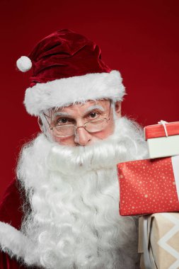 Şapkalı Noel Baba 'nın portresi. Elinde hediye kutuları ve kırmızı arka plandaki kameraya bakıyor.