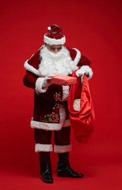 Kırmızı kostümlü ve güneş gözlüklü Noel Baba kırmızı arka planda duruyor ve hediyeleri torbaya koyuyor.