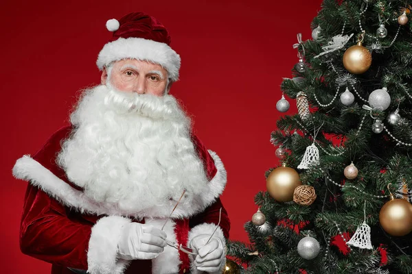 Portrait of Santa Claus with white beard and in red costume posing at camera near the Christmas tree on red background