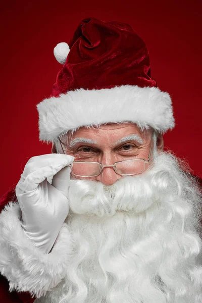 圣诞老人头戴帽子 留着白胡子 戴着眼镜 在红色背景下看着相机的特写镜头 — 图库照片