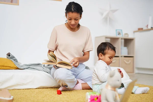 Young mother sitting on the floor and reading book to little child while playing with toys