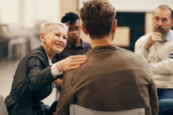 Smiling mature woman with short blond hair talking to the young man who sitting on the chair back to the camera during therapy class