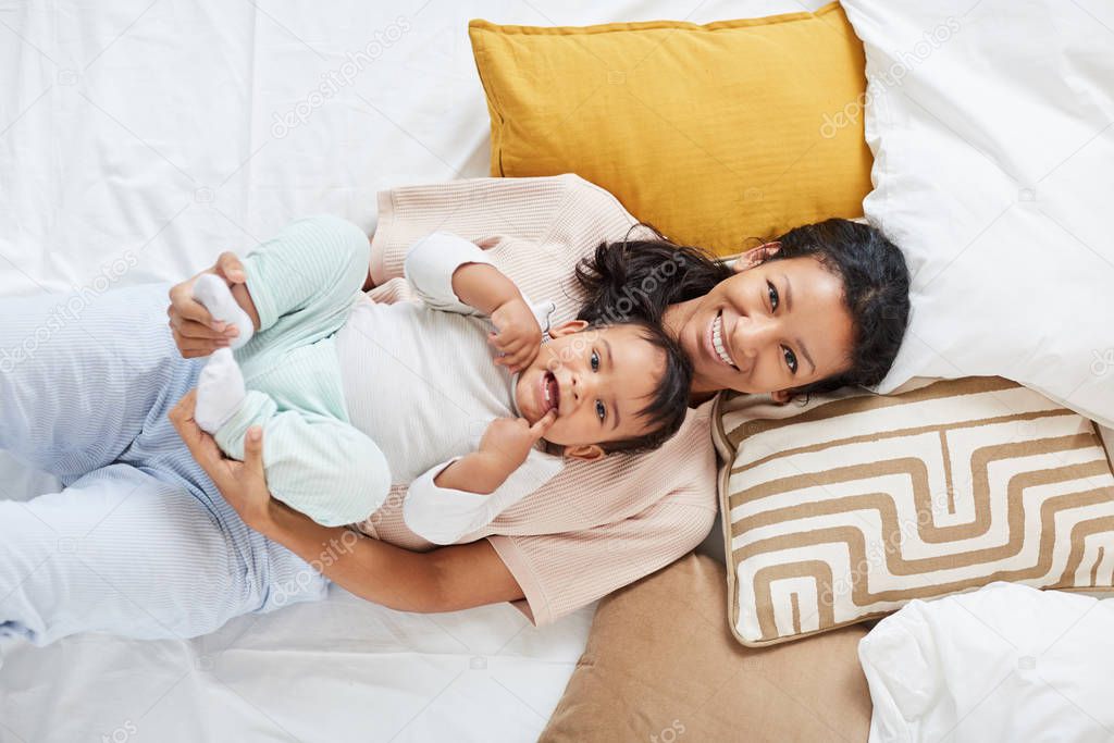 Portrait of young happy mother smiling at camera while resting on the bed with baby