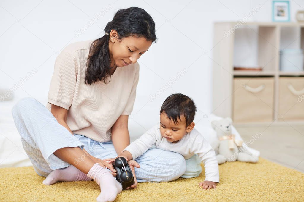 Young mother in casual clothing sitting on the floor and showing the clock to little child