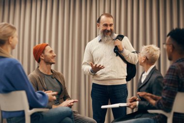 Senior man with beard standing and presenting idea to people sitting on chairs and listening 