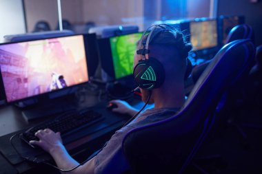 Rear view of young gamer wearing gaming headphones with backlight and playing in computer video game on computer in dark computer class clipart