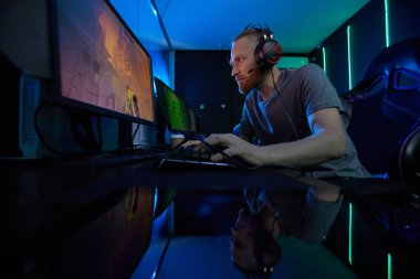 Kulaklıklı genç sakallı adam oyun sandalyesinde oturuyor ve bilgisayar oyununda oynuyor karanlık odada. 