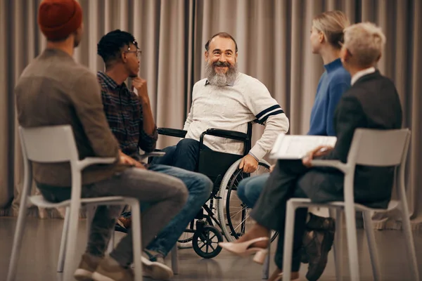 Happy senior disabled man sitting in wheelchair and talking to the young people who sitting on chairs in circle during therapy lesson