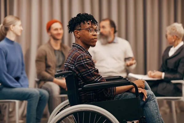 Young African disabled man sitting in wheelchair with people talking to each other in the background