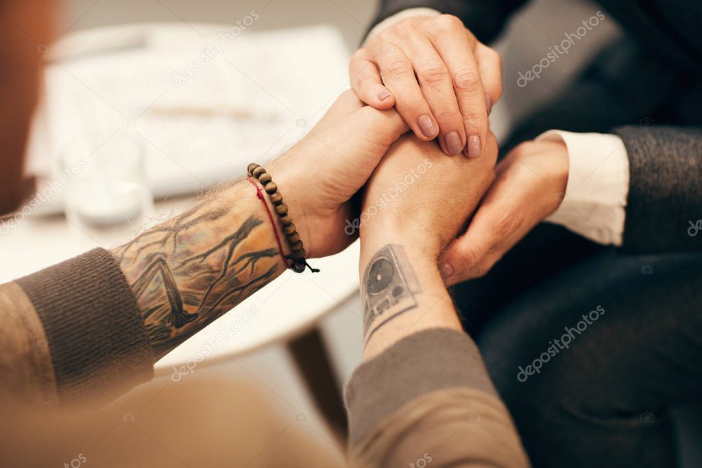 Close-up of mature woman holding hands of young man and supporting during visit to psychologist