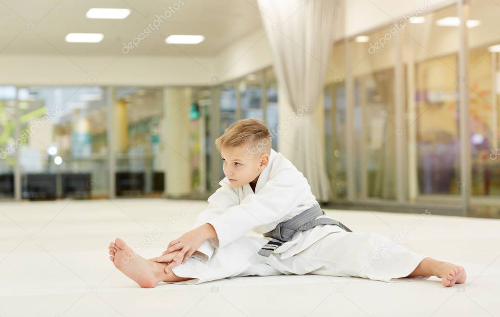 Little karateka in kimono doing stretching exercises on the floor in gym