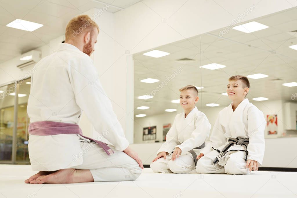 Rear view of young coach in kimono sitting on the floor and greeting two boys looking and smiling in the gym