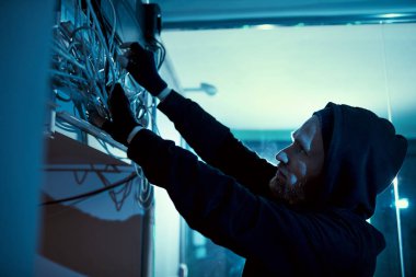 Siyah kapüşonlu gömlekli elektrikçi elinde kabloyla odadaki elektriği tamir etmeye çalışıyor.