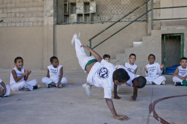 Rio de Janeiro, Brezilya - 9 Ağustos 2016: Brezilya 'da Capoeira sınıfındaki çocuklar Berimbau oynayan bir öğretmenle arka planda bir gecekondu mahallesindeki açık hava spor sahasında arkadaşlarıyla çember oluşturuyorlar.