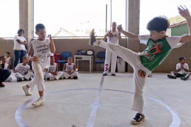 Rio de Janeiro, Brazil - August 9, 2016: Kids at a Brazilian capoeira class with a teacher playing a berimbau instrument in the background at an outdoor sports venue in a shantytown clipart