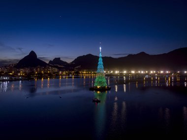 Rio de Janeiro gölünün ortasındaki yüzen Noel ağacının ışıkları gün batımında yeşil ve mavi renkte.