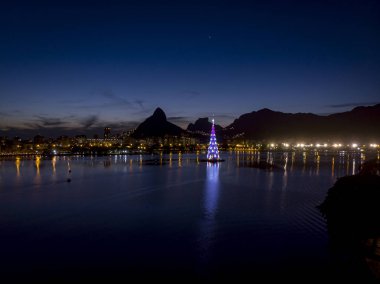 Gün batımında Rio de Janeiro gölünün ortasındaki yüzen Noel ağacının ışıkları mavi ve mor renkte.