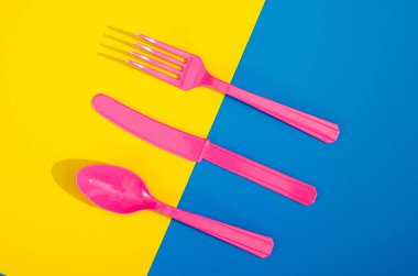 Renkli arka planda duran pembe plastik tek kullanımlık çatalların, kaşıkların ve bıçakların derisi yüzülmüş. Yaratıcı üst görünüm deseni.