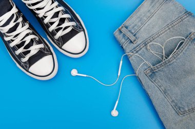 Mavi ve beyaz spor ayakkabılar, kablosuz kulaklıklar ve mavi arka planda duran kot pantolonlar. Üst Manzara. Flatlay. Kopyalama Merkezi.