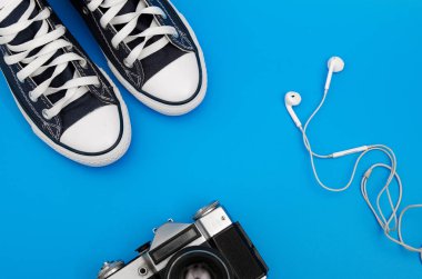 Mavi ve beyaz spor ayakkabılar, kablosuz kulaklıklar ve mavi arka planda bir kamera var. Üst Manzara. Flatlay. Kopyalama Merkezi.