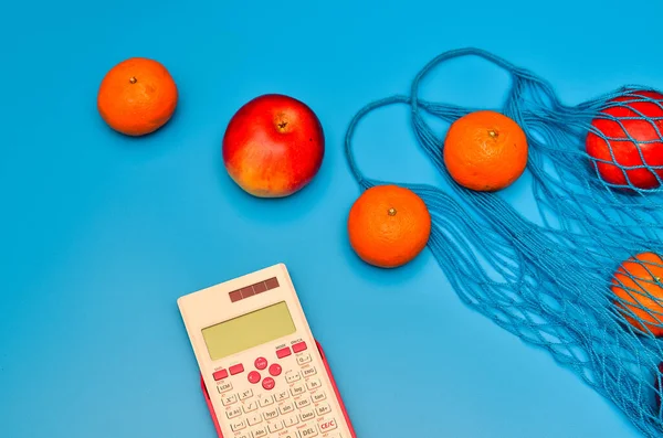 Pembe hesap makinesi ve sağlıklı yiyeceklerle dolu bir alışveriş çantası. Moda mavi arka planda farklı meyvelerden yapılmış özel tasarım el çantasının yemek fotoğrafçılığı. 2020 yılının klasik mavi rengi