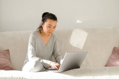 Gri gömlek ve pantolon giymiş Asyalı bir kadın. Kanepede otururken evden çalışan genç Asyalı kadın. Dizüstü bilgisayar kullanan Koreli kız. Chihuahua köpeğini tutan kız.