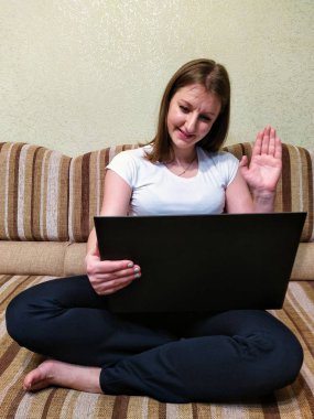 Gülümseyen genç kadın kanepede oturur, video görüşmesi yapar ve evde el sallar.