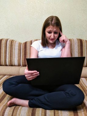 Üzgün genç bir kadın kanepede oturur ve evdeki bilgisayarında görüntülü arama yapar.