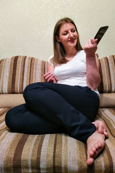 Χαμογελαστή Νεαρή Γυναίκα Κάθεται Στον Καναπέ Και Χρησιμοποιήσετε Smartphone Στο — Φωτογραφία Αρχείου