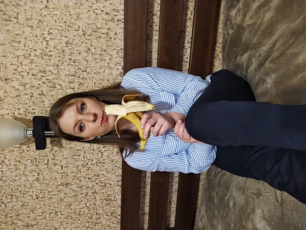 European woman sit on bed and eat banana in bedroom at home