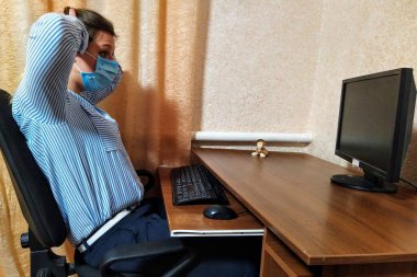 Ürkmüş Avrupalı maskeli genç bir kadın masada oturup evdeki bilgisayarda çalışıyor.