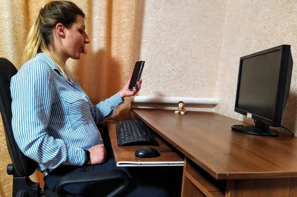 欧洲的年轻女子坐在桌旁 一边打电话一边在家里的智能手机上伸出舌头 — 图库照片