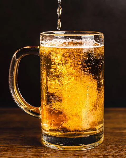 Cold beer with foam in a mug, on a wooden table and a dark background with blank space for a logo or text. Stock Photo mug of cold foamy beer close-up.