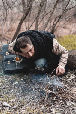 Kendini ısıtan, küçük bir ateşin başında oturan, doğada yaşayan bir adamın resmi..