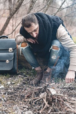 Kendini ısıtan, küçük bir ateşin başında oturan, doğada yaşayan bir adamın resmi..